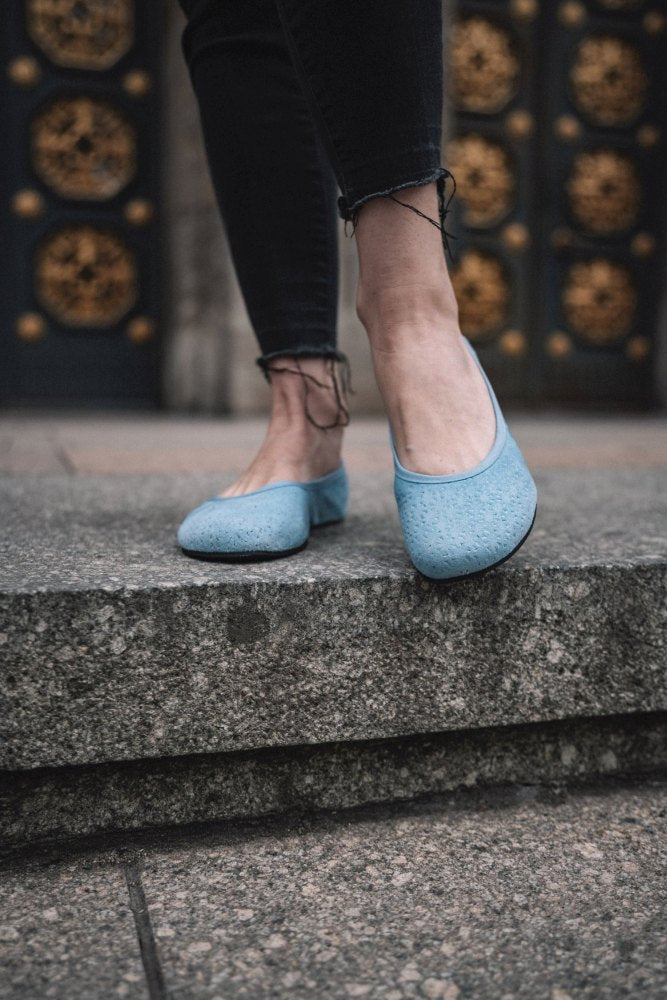 Auf einer Steinplattform steht eine Person in ausgefransten schwarzen Jeans und präsentiert ein Paar TWIST Light Blue Slip-On-Schuhe von ZAQQ, ideal für flexible Sommerlooks. Die verzierte Tür mit goldenen und schwarzen Details bietet einen eleganten Hintergrund.