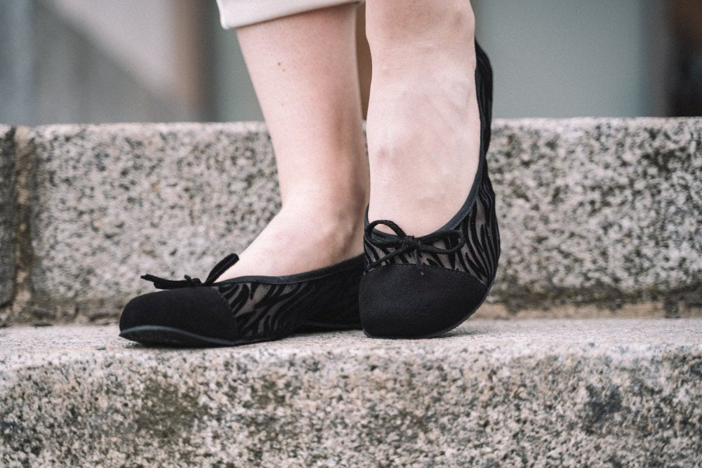 Nahaufnahme einer Person, die ZAQQs TWIST Sling Black-Schuhe mit schwarzem Zebramuster und flexibler Sohle trägt und auf einer Steintreppe steht. Sie trägt helle Hosen und genießt den Komfort dieses stylischen Schuhwerks.