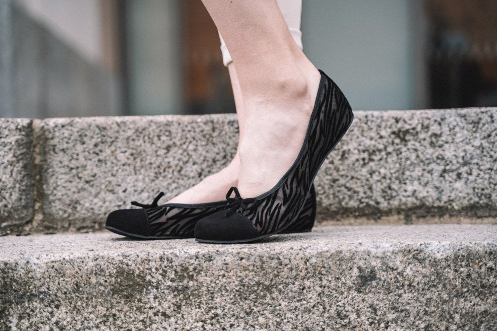 Nahaufnahme einer Person, die die TWIST Sling Black Flats mit Zebramuster von ZAQQ trägt und auf Steinstufen steht. Diese bequemen Schuhe haben kleine Schleifen auf der Vorderseite und eine flexible Sohle für zusätzlichen Komfort.