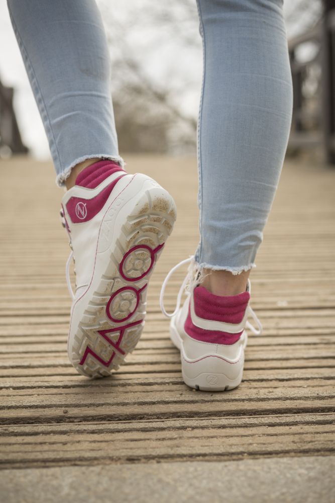 Eine Person in hellblauer Jeans und pinken Socken schlendert einen Holzweg entlang. Sie trägt weiße Sneakers mit pinken Akzenten, die an den ZAQQ VAQATION Pink Waterproof-Stil erinnern. Der unscharfe Hintergrund lässt Bewegung erahnen.