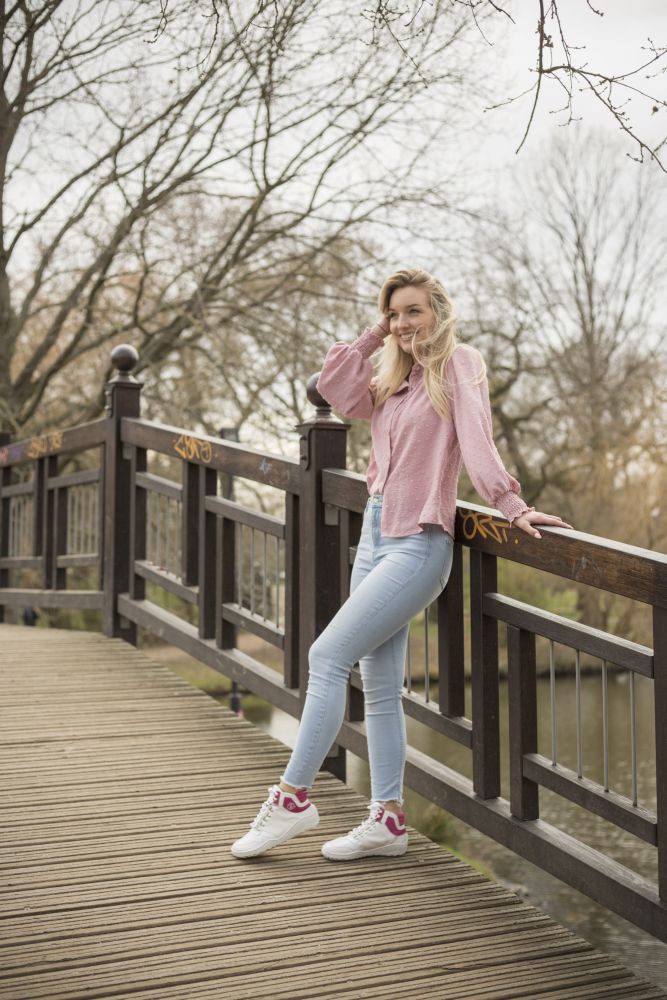 Eine Person, die einen ZAQQ VAQATION Pink Waterproof Pullover und hellblaue Jeans trägt, steht auf einer Holzbrücke und lehnt sich an das Geländer. Im Hintergrund sind kahle Bäume und ein ruhiger Fluss zu sehen. Die Person lächelt, hat langes, welliges Haar und trägt bequeme Barfußschuhe.